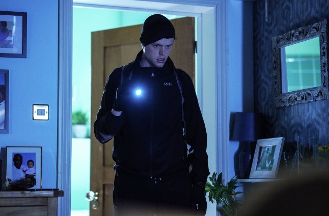 Oscar walks through the Trueman house in black clothes and holding a torch in EastEnders