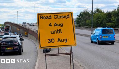 A view of the flyover with cars driving over it. Several drive to the side of it. A yellow sign in the middle says "Road closed - 4 Aug until 30 Aug".