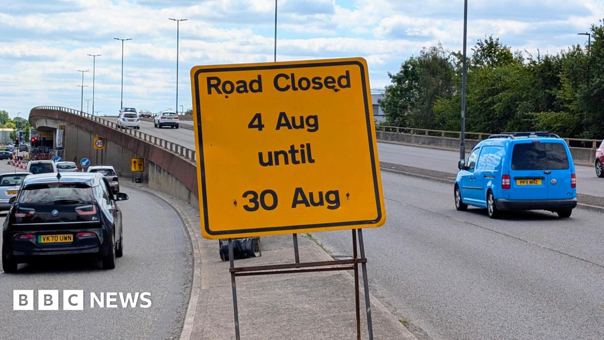 A view of the flyover with cars driving over it. Several drive to the side of it. A yellow sign in the middle says "Road closed - 4 Aug until 30 Aug".