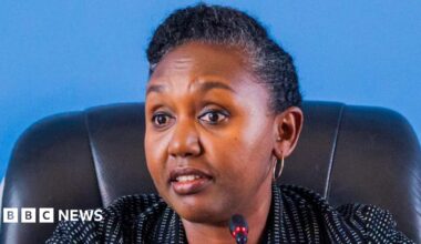 Rwanda government spokesperson Yolande Makole sits on a black chair before a microphone. She is wearing a white dotted black blazer.