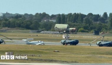 Small white plane on runway surrounded by large vehicles with small crane arms on them.