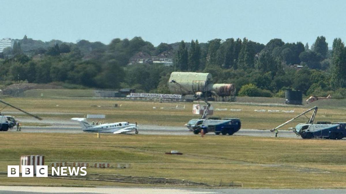 Small white plane on runway surrounded by large vehicles with small crane arms on them.