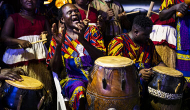 A group of Black drummers dressed in traditional garb clap and drum together.