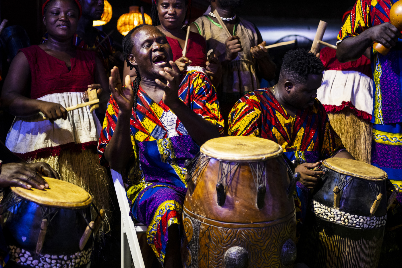 A group of Black drummers dressed in traditional garb clap and drum together.