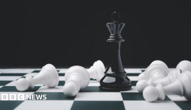 On a black and white chess board, a black king chess piece stands upright. It is surrounded by toppled white pawns.