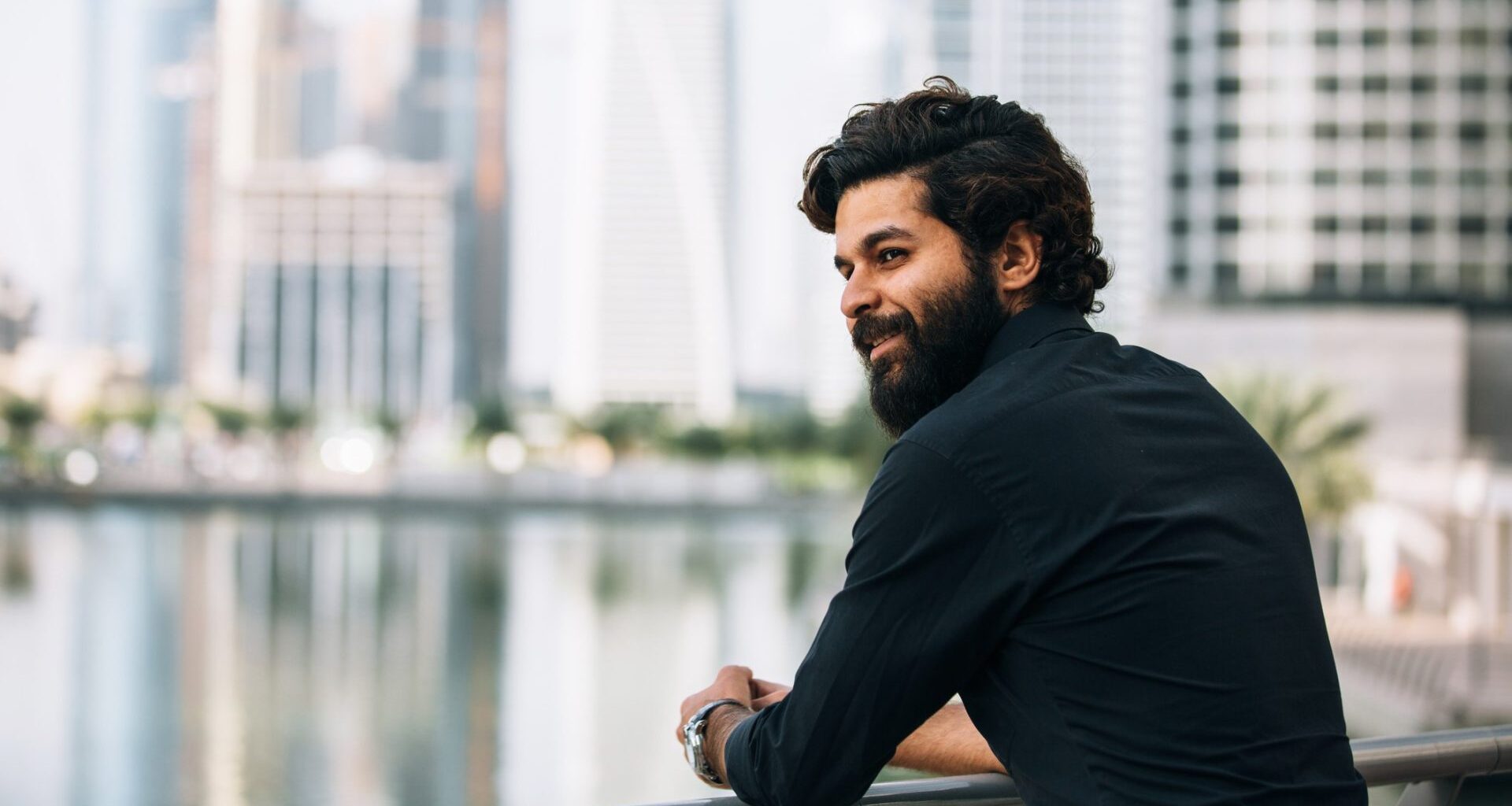 Young man with beard standing outside with a city backdrop, showing quiet wealth and luxury.
