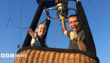 Doug and a woman in a balloon basket. He has his thumb up and his other hand on the balloon burner. His female passenger is smiling and waving at the camera