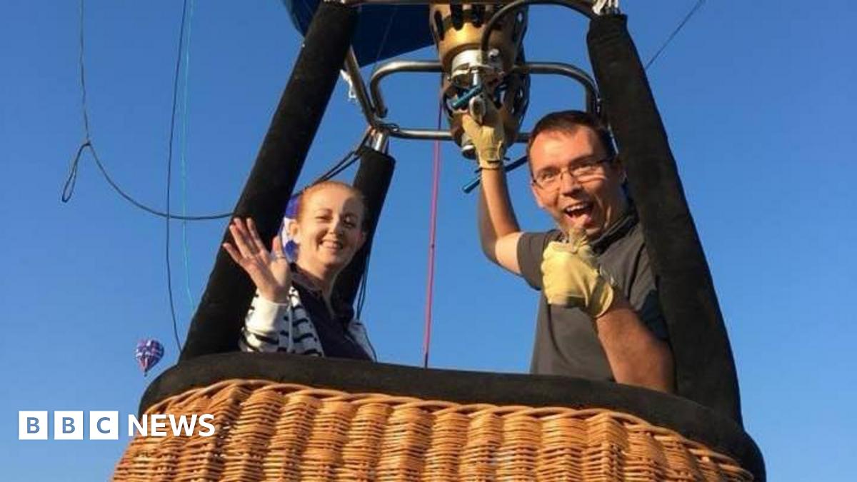 Doug and a woman in a balloon basket. He has his thumb up and his other hand on the balloon burner. His female passenger is smiling and waving at the camera