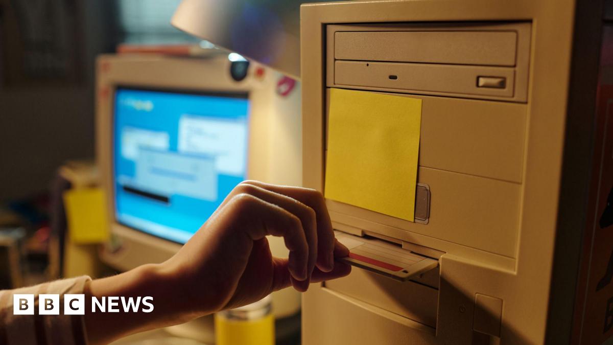 Hand inserting floppy disk into vintage computer with background showing another computer monitor displaying a blue screen.