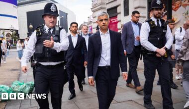 Sadiq Khan, a middle-aged man wearing a suit, walking on a busy shopping street with male police officers walking either side of him.