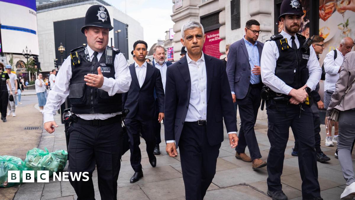 Sadiq Khan, a middle-aged man wearing a suit, walking on a busy shopping street with male police officers walking either side of him.