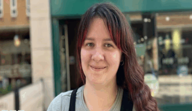 Sian has brown hair with red highlights on her fringe is wearing a grey blue top and black dungarees over it.