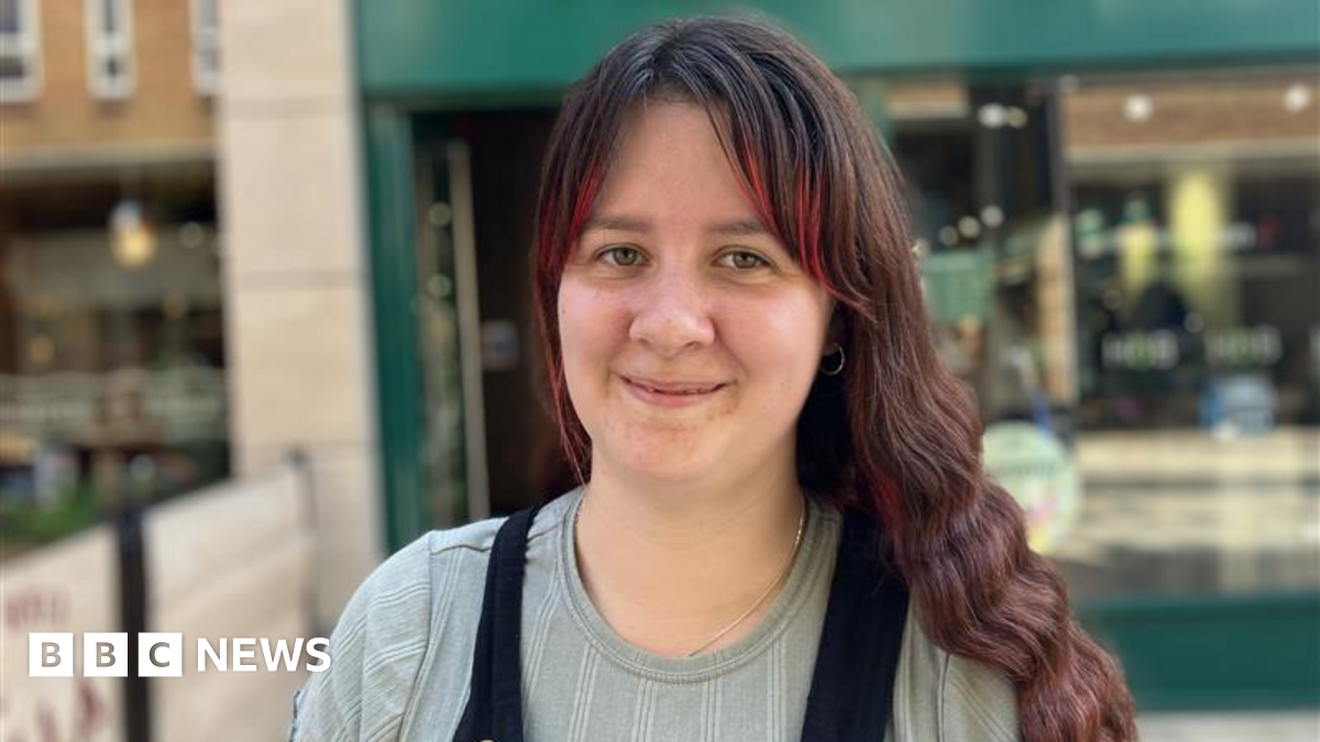 Sian has brown hair with red highlights on her fringe is wearing a grey blue top and black dungarees over it.