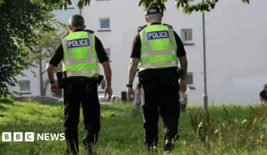 Two police officers with their back to the camera. Both are wearing dark clothing and boots with hi-viz jackets over the top with the word police on the back in white writing on a blue background. The officer on the right is wearing a cap. They are walking through long green grass towards a white block of flats with small windows in the side.