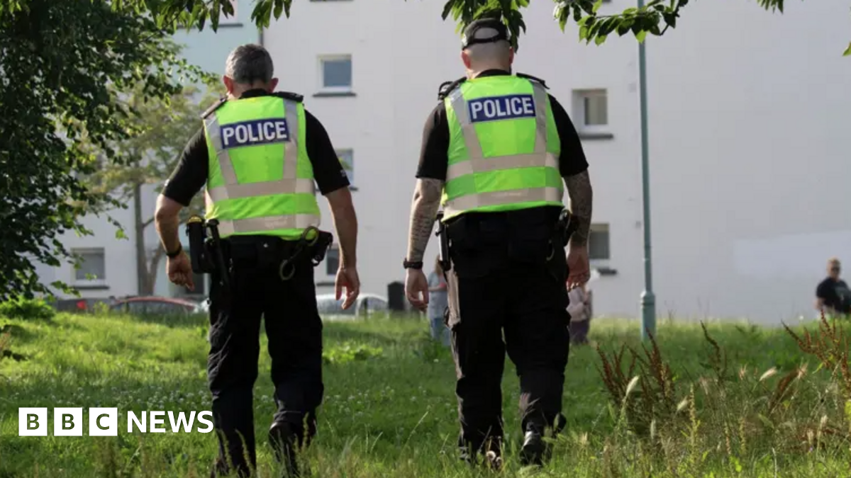 Two police officers with their back to the camera. Both are wearing dark clothing and boots with hi-viz jackets over the top with the word police on the back in white writing on a blue background. The officer on the right is wearing a cap. They are walking through long green grass towards a white block of flats with small windows in the side.