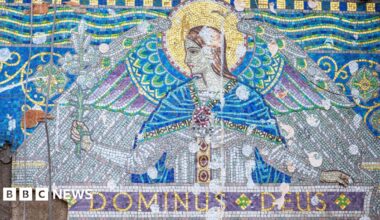 A mosaic frieze consisting of decorative Venetian glass tiles in white, purple, gold and blue, depicting an angel with the Latin 'Dominus Deus' running a long a banner underneath. The display is placed uncovered on a partially demolished wall.