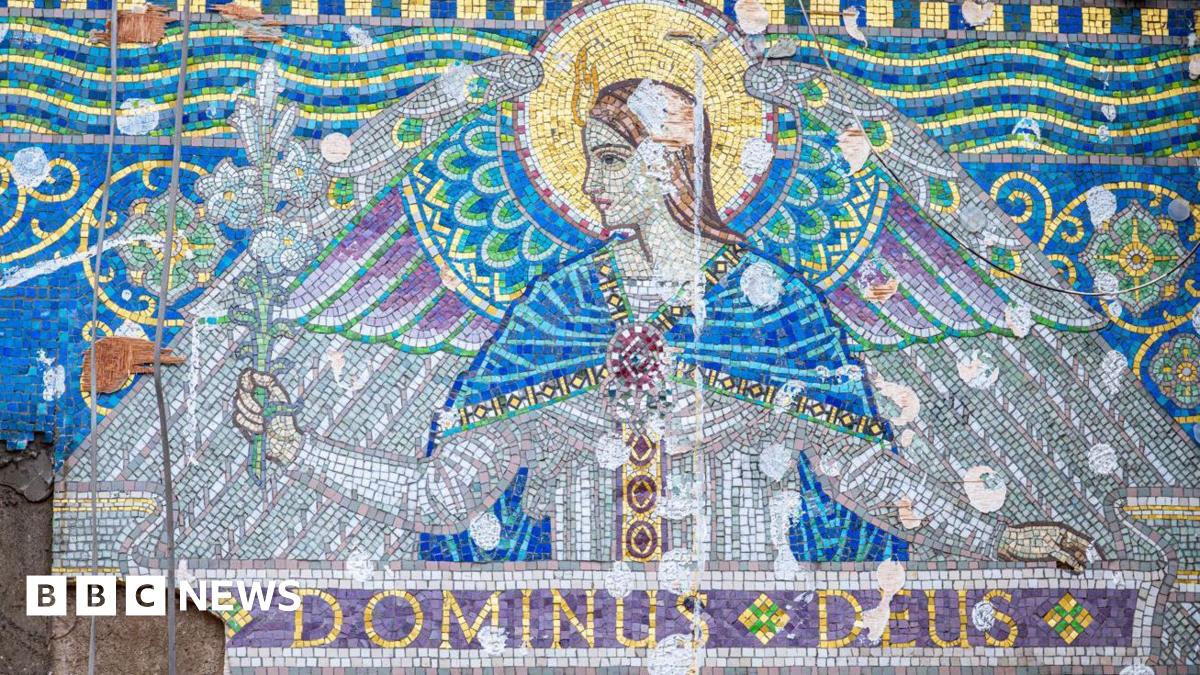 A mosaic frieze consisting of decorative Venetian glass tiles in white, purple, gold and blue, depicting an angel with the Latin 'Dominus Deus' running a long a banner underneath. The display is placed uncovered on a partially demolished wall.