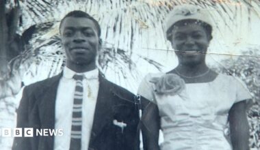 Annetta Stanley is looking directly at the camera.  She has long, braided, hair and is wearing a scarf, tied in a bow, on her head.  She is wearing a white shirt and a gold coloured necklace.