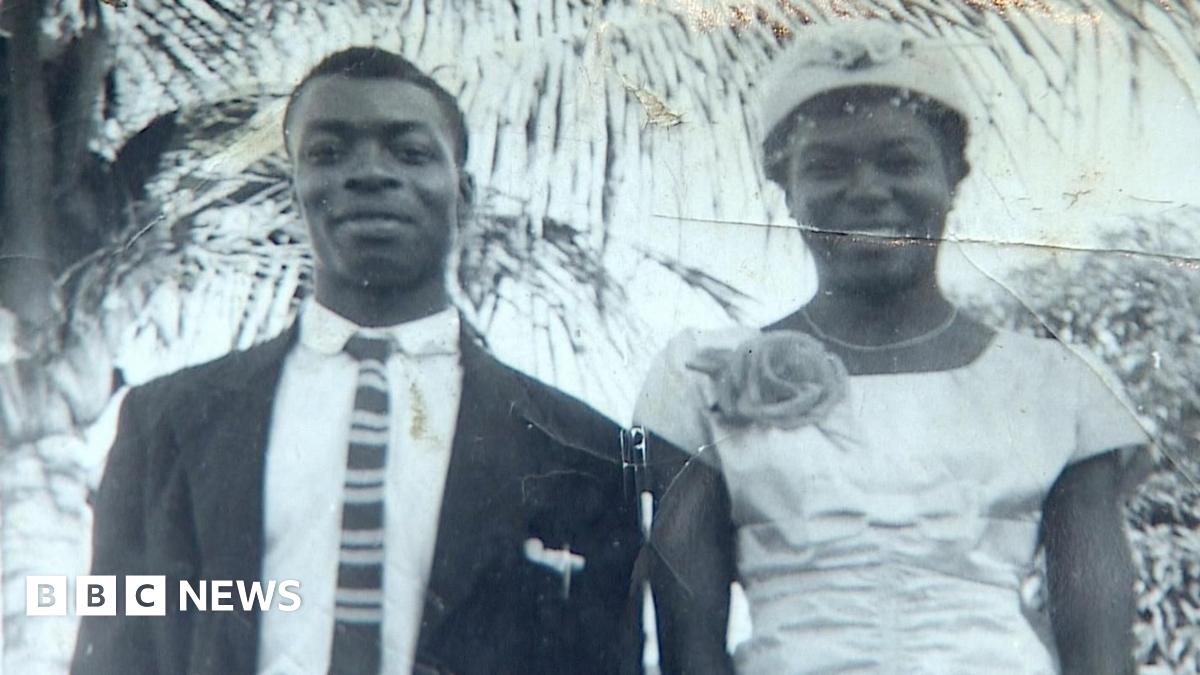 Annetta Stanley is looking directly at the camera.  She has long, braided, hair and is wearing a scarf, tied in a bow, on her head.  She is wearing a white shirt and a gold coloured necklace.