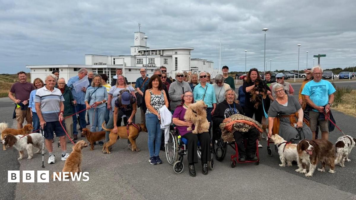 About 30 people, many with dogs, gather on Birkdale Common to protest plans to allow the R&A to take over a section of the public land ahead of the 2026 Open golf championships there. Three people in wheelchairs sit in the foreground, in front of the standing protesters.