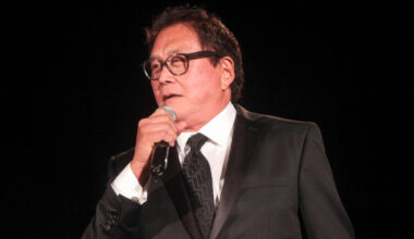Robert Kiyosaki in a suit talking at an event, holding a microphone