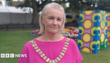 Lord Mayor Tracy Kelly is wearing pink and a gold chain of office. She has short blond hair. She's standing with grass and trees behind her. Some colourful stands have been erected.