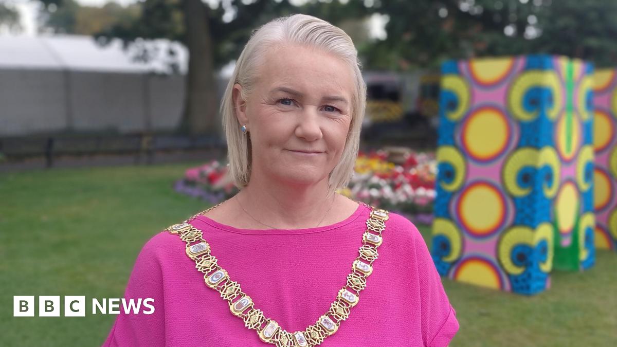 Lord Mayor Tracy Kelly is wearing pink and a gold chain of office. She has short blond hair. She's standing with grass and trees behind her. Some colourful stands have been erected.