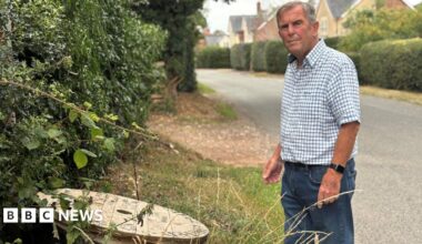 A man, wearing is a blue and white check shirt and blue jeans, is standing on the roadside looking at a wooden reel of cables next to the hedge.