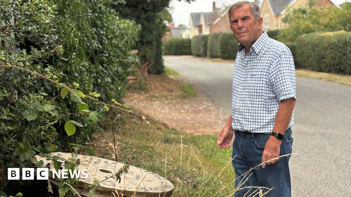 A man, wearing is a blue and white check shirt and blue jeans, is standing on the roadside looking at a wooden reel of cables next to the hedge.