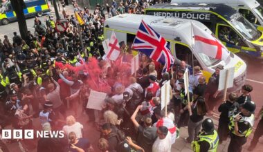 Anti-immigrations protesters waving St George and union jack flags are on one side of a line of police officers on a street. One person is holding a red flare, leaving red smoke above some people's heads. On the other side are counter protesters.  There are three police vans to the side of them; the middle one says Heddlu Police and is black.