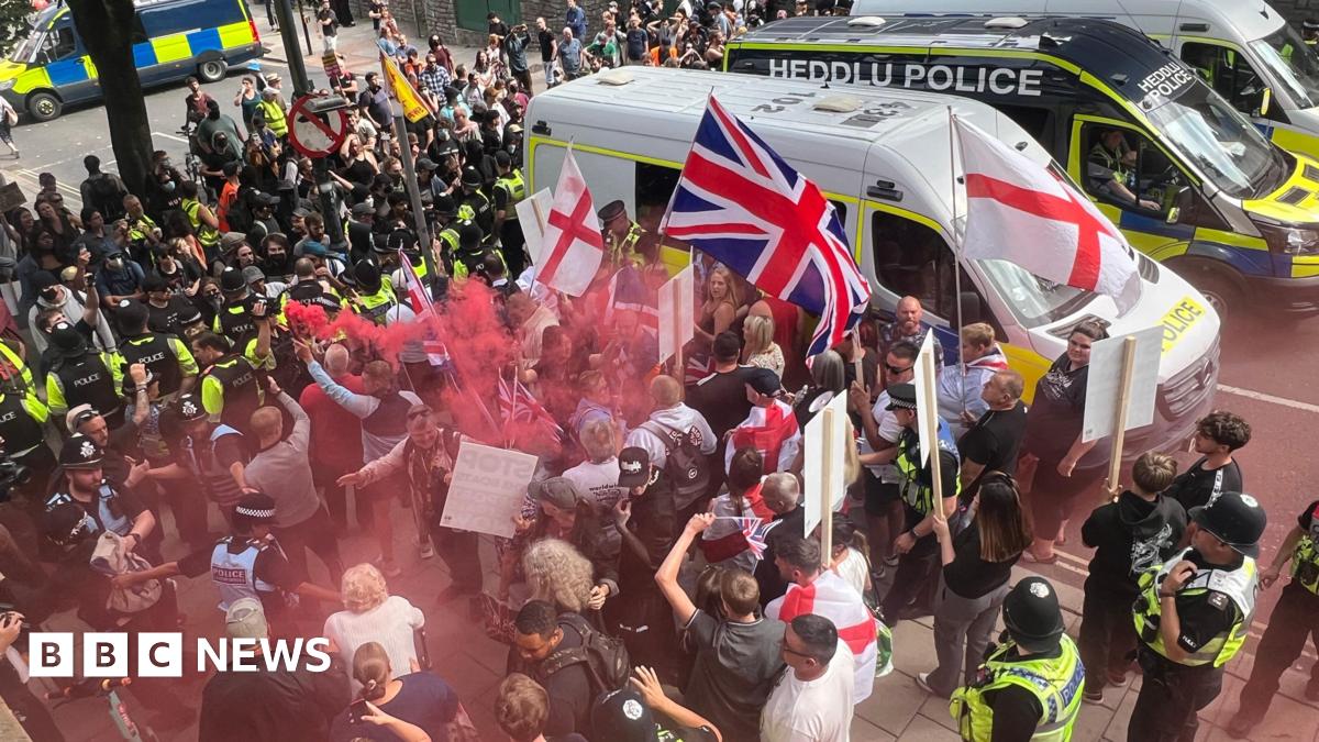 Anti-immigrations protesters waving St George and union jack flags are on one side of a line of police officers on a street. One person is holding a red flare, leaving red smoke above some people's heads. On the other side are counter protesters.  There are three police vans to the side of them; the middle one says Heddlu Police and is black.