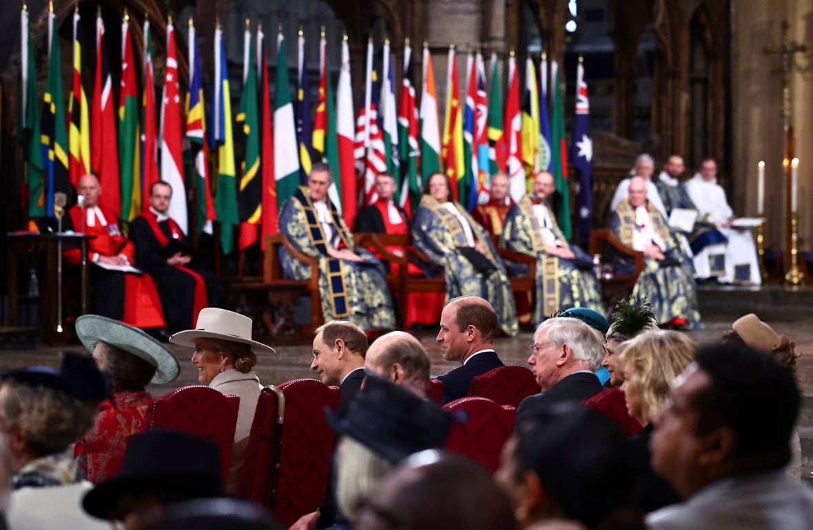 A row of flags stand in the background as people sit in a large abbey.