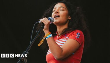 A young woman with curly dark hair closes her eyes as she sings into a microphone. She is wearing a red top with white stripes on it and has two festival wristbands around her wrist.