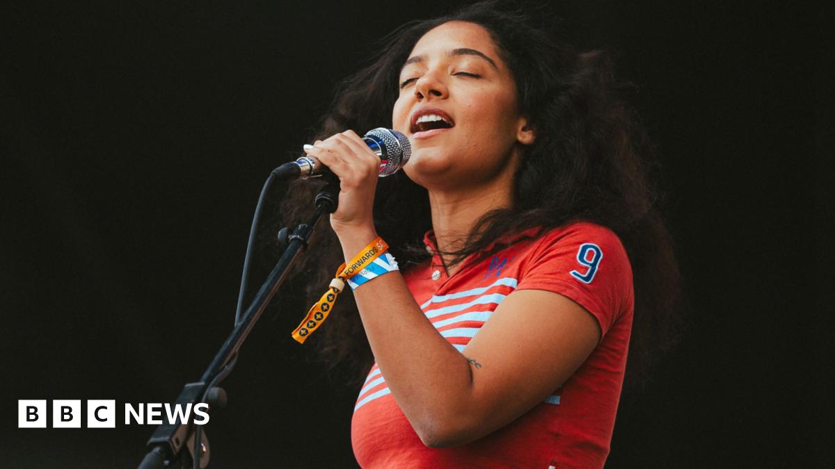A young woman with curly dark hair closes her eyes as she sings into a microphone. She is wearing a red top with white stripes on it and has two festival wristbands around her wrist.