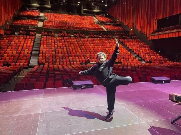 Dakota, in all black, standing on one leg with her left arm raise at a stage. Hundreds of red chairs can be seen in the background