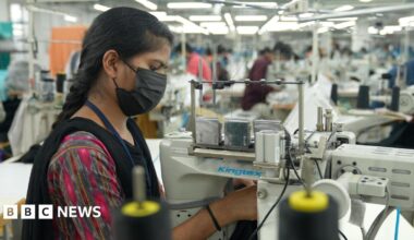 A young woman, wearing a multi-coloured dress with a mask, intently working on a sewing machine at a factory in southern India.