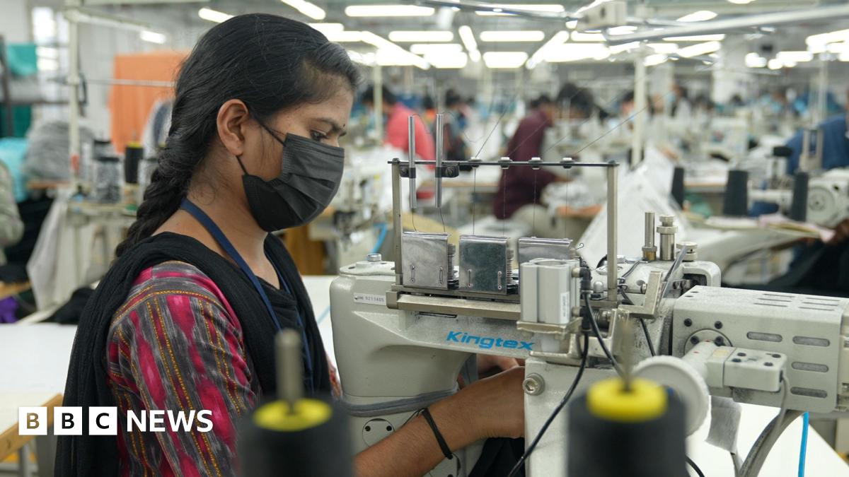 A young woman, wearing a multi-coloured dress with a mask, intently working on a sewing machine at a factory in southern India.