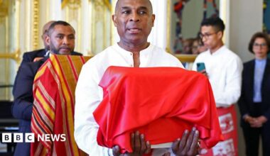 A Malagasy official holds a box covered in a red cloth during a ceremony at the French culture ministry to return the skull of King Toera. Behind him are two other officials holding boxes - one is covered in a red, yellow and black stripped material. Out of focus to the right France's Culture Minister Rachida Dati looks on - Tuesday 26 August 2025.