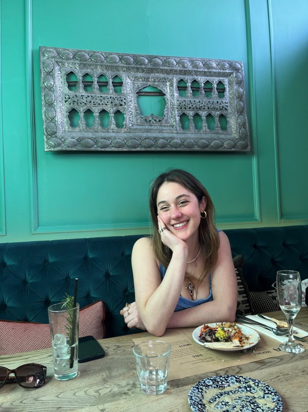 A young woman smiling while sitting at a table in a restaurant, with a decorative wall panel behind her. She has a plate of food in front of her, alongside glasses and a phone on the table.