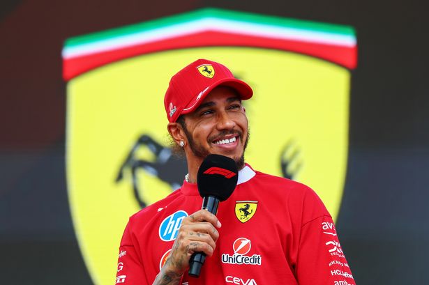 Lewis Hamilton smiles as he speaks into a microphone in front of a giant Ferrari crest