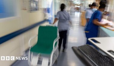 A blurred image of staff working in a hospital corridor wearing blue scrubs. One staff member is pulling a green wheelchair.