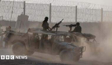 Members of the Israel Defence Force stand in two open-topped military vehicles near a fenced border, which is topped with barbed wire. Each vehicle has a large gun extending from its roof, manned by a person in a helmet and camoflague uniform.