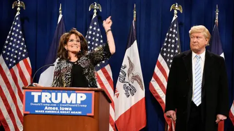 NBCU Photobank Tina Fey stands at a lectern with a "Trump" sign on the front as she plays Sarah Palin, US flags i a row behind her. Darrell Hammond as Donald Trump is to her left, giving her the "side eye" look.