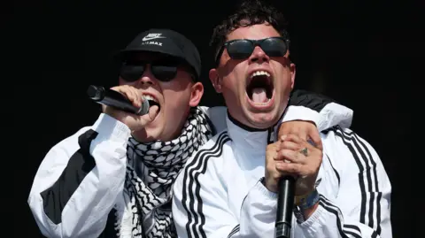 Andy Rain/EPA/Shutterstock Kneecap's Mo Chara and his bandmate Móglaí Bap perform on the West Holts stage during day four of the Glastonbury Festival.  They are both wearing dark sunglasses and black and white track suit or sports jackets.   Mo Chara has his arm around Móglaí Bap's shoulder. 