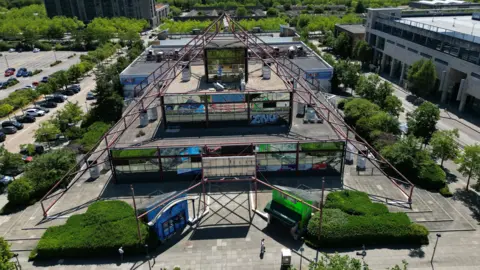 BBC A view of the Point from above via a drone. It is a glass pyramid type structure with red metal supporting beams.