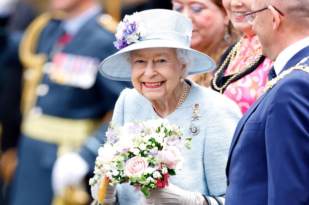 Photo of Queen Elizabeth II, wearing a light blue hat and coat, smiling at the camera and holding a bouquet of flowers outside the Palace of Holyroodhouse, Edinburgh, 2022