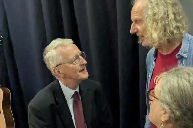Secretary of State for NI Hilary Benn in a dark i sitting at the edge of a small stage speaking to RARE founder Colum Sands in a denium shirt and Carlingford Lough Keepers found Fionnuala McKinley in a multi-coloured top and glasses, both of them are standing. 