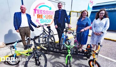 (L-R) Councillor Peter Gavigan, SUEZ Regional Director Steve Patterson, Councillor Lisa Evans and Amanda Padfield, director of public sector development at SUEZ. All four are standing in front of a small bike rack which is supporting seven children's bicycles. A sign on the wall marks the site as the Teesside Reuse Shop.
