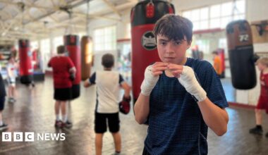 A group of children training in a boxing gym with several punching bags hanging from the ceiling. In the foreground, Atti is wearing hand wraps is either preparing for or taking a break from training. In the background, others are actively engaged in boxing exercises