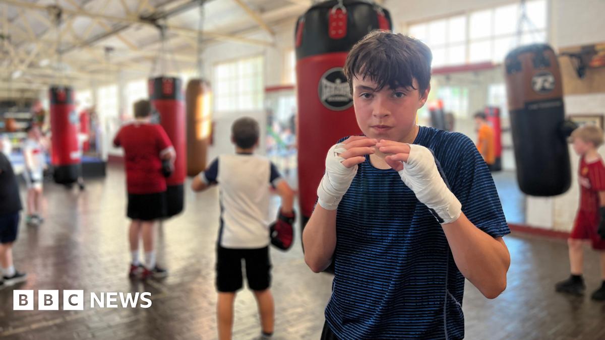 A group of children training in a boxing gym with several punching bags hanging from the ceiling. In the foreground, Atti is wearing hand wraps is either preparing for or taking a break from training. In the background, others are actively engaged in boxing exercises